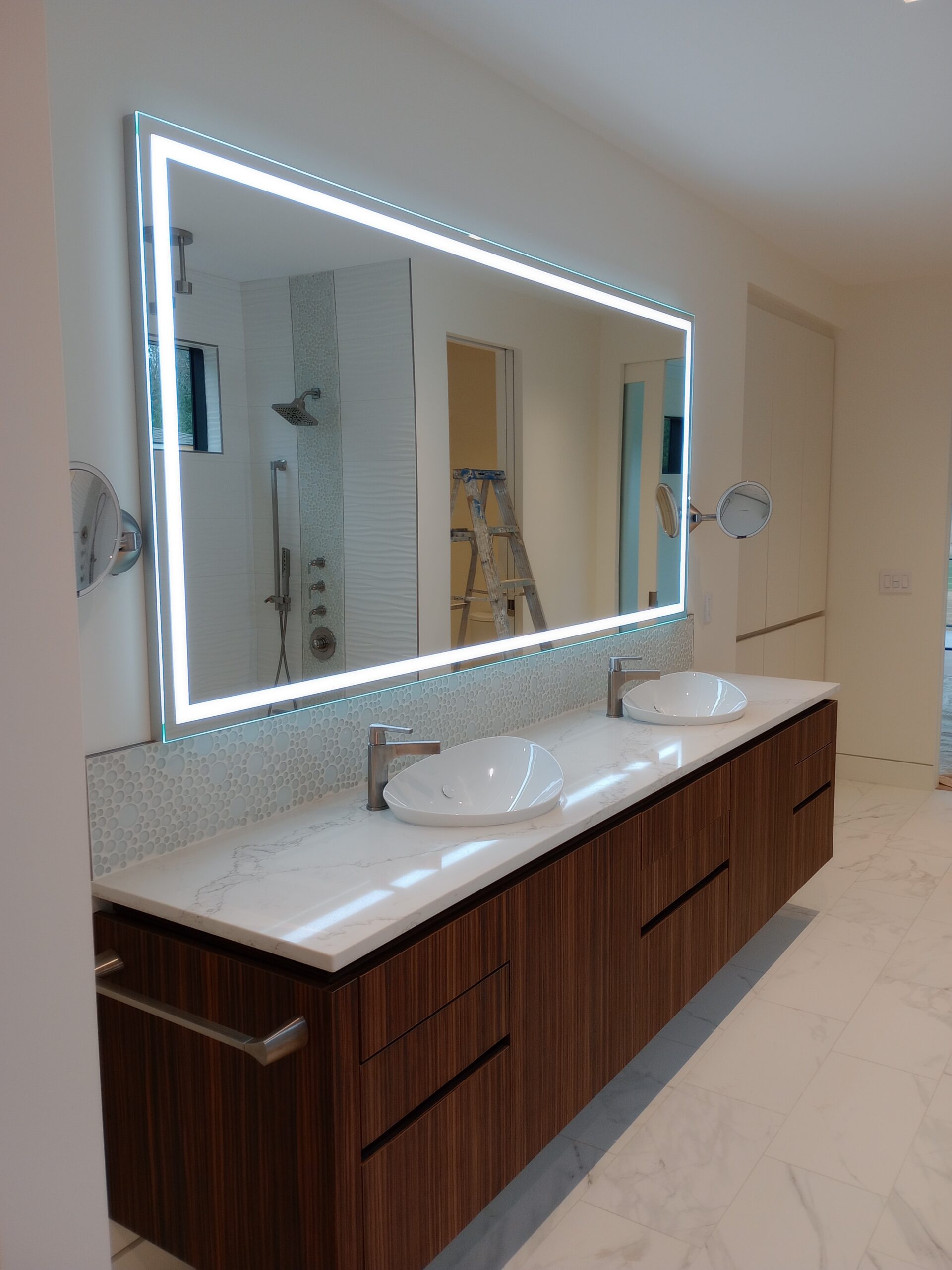 Painted bathroom walls beside a double vanity and illuminated mirror, with a ladder reflected in the mirror